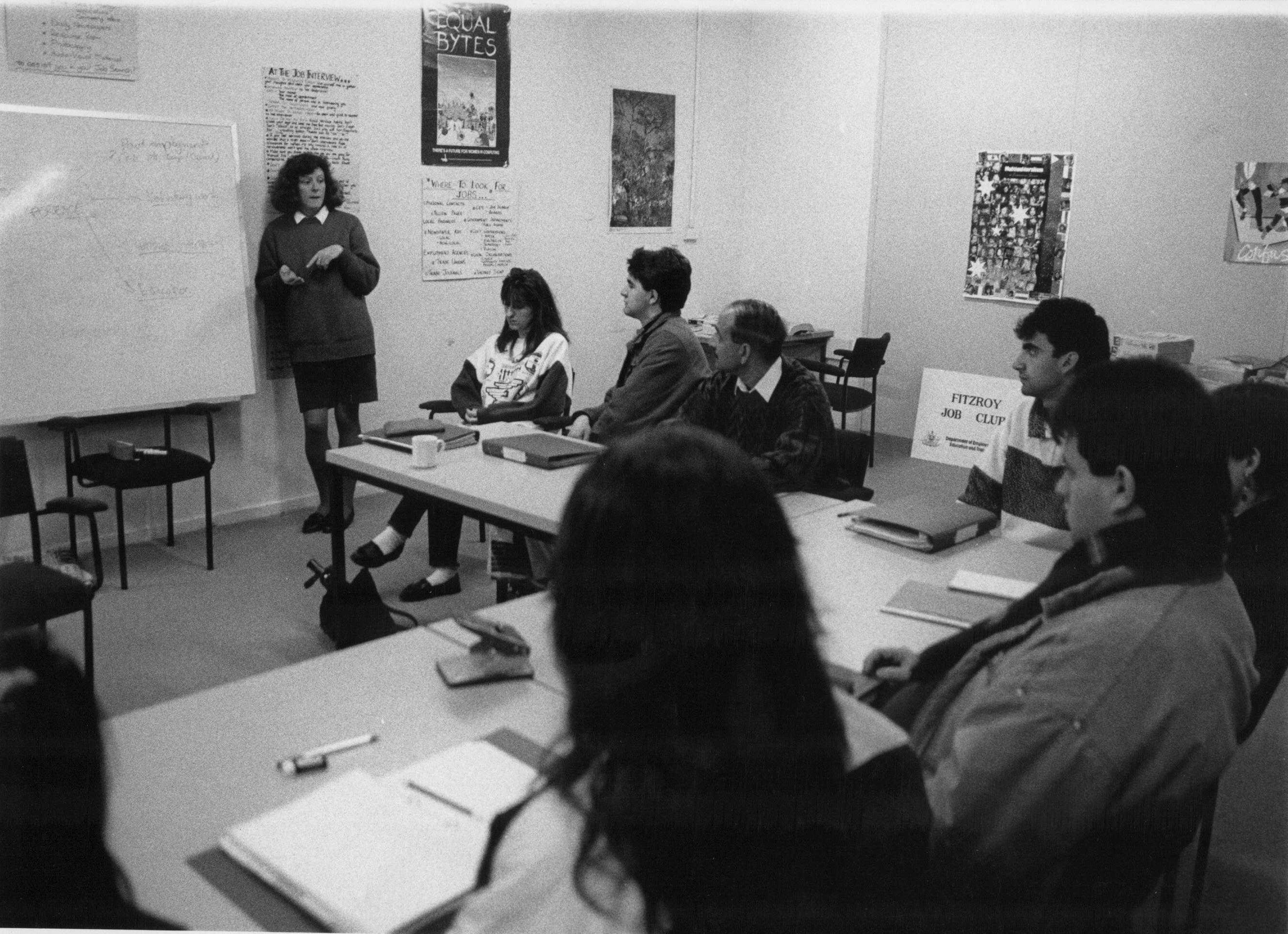 This is a black and white photo of an employment officer teaching job seekers. There are about 10 people listening to the employment officer. She is standing in front of a whiteboard.
